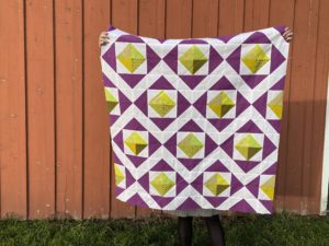 diamond quilt with faceted yellow-green center diamonds, a completed quilt top held up against a barn with a grey sweater dress and black tights sticking out the bottom.