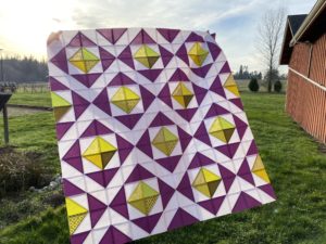 diamond quilt with faceted yellow-green center diamonds, a completed quilt top held against the sun for a stained glass effect. The bottom left corner of the quilt moves closer through the depth of field and there is a sun-flare in the upper left of the image