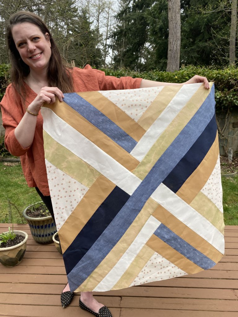 A quilt top rendered in shades of brown, blue and green. The modified log cabin design gives the impression of a woven maypole. The quilt is held by a long-haired brunette femme holds a completed quilt top. She is in a coral-colored linen top on a deck with a few pots.