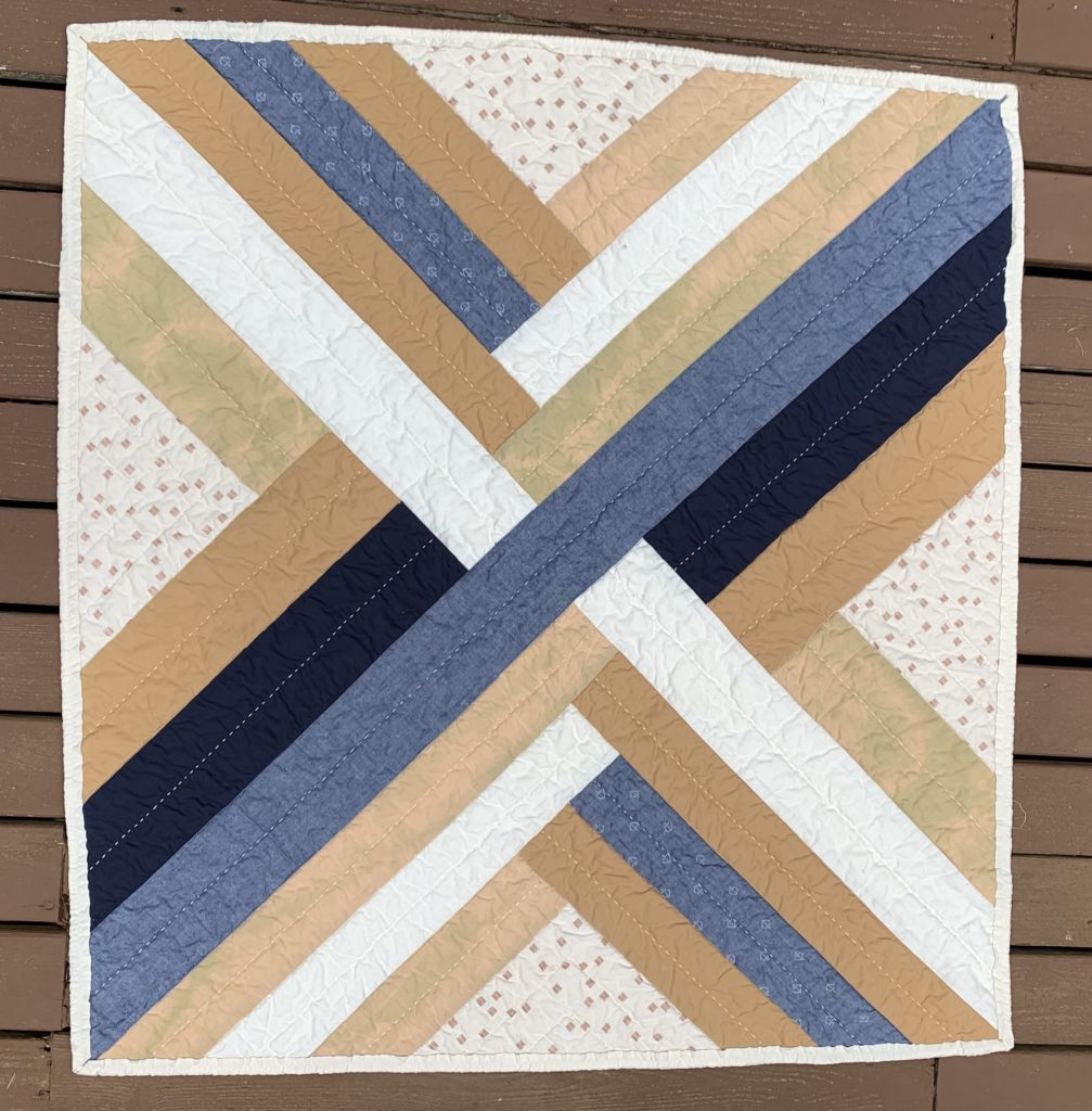 maypole quilt: an overhead view of a nearly-square quilt on a wooden deck. The effect is of woven ribbons. The colors are based on those seen in the Pacific Northwest. There are two shades of medium brown, metallic speckled neutral, chambray (some with anchors), navy and ice-dyed neutral fabric
