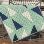 pescadero: a baby-sized quilt lays draped over a railing. The background is in mint, and there are white, admiral blue and navy blue half-square triangles and half-rectangle triangles. The binding is steel essex linen. The sun is shining, highlighting the wavy-stitched diagonal quilting on cotton batting.