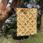 spectrum: a quilt using shades of yellow, green, orange and brown rectangles to create a cascading, tumbling effect. the quilt is held by someone wearing green boots and nail polish in front of a madrona tree during early pandemic