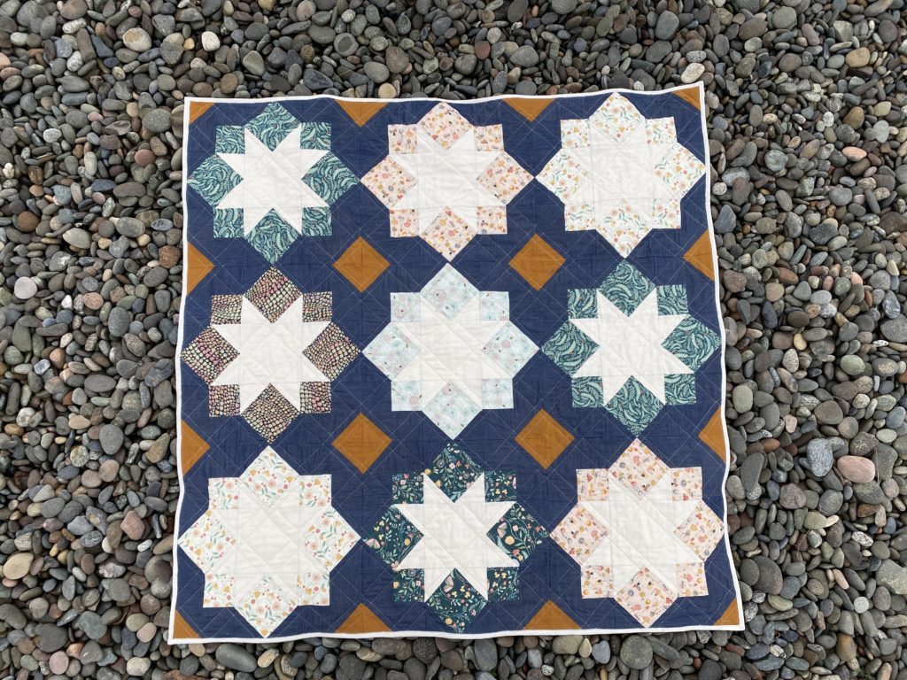 stellar mosaic: a square quilt lies on the rocks at driftwood beach in Keystone, WA. the quilt has a 3x3 grid of stars outlined by rotated squares. the printed fabric line is called "frond of you," and the chambray is from District Fabrics in Pt. Townsend, WA