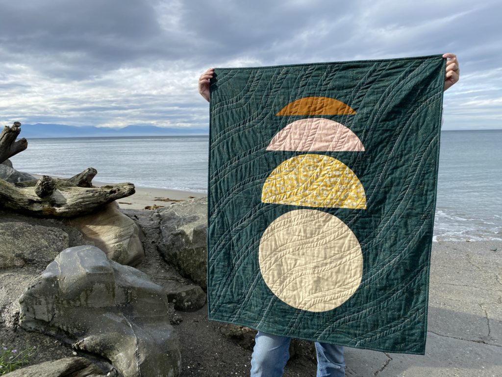clava: a small quilt held by a tall man at Hastie Lake Beach on Whidbey Island, WA. The quilt has a speckled pine green background and binding. a champagne-colored circle has progressively smaller semicircle stones placed on top. The circle slices are yellow metallic stars, pink metallic stars and cinnamon brown. the piece is machine-quilted using a scalloped decorative stitch and straight stitching in glow-in-the-dark thread.
