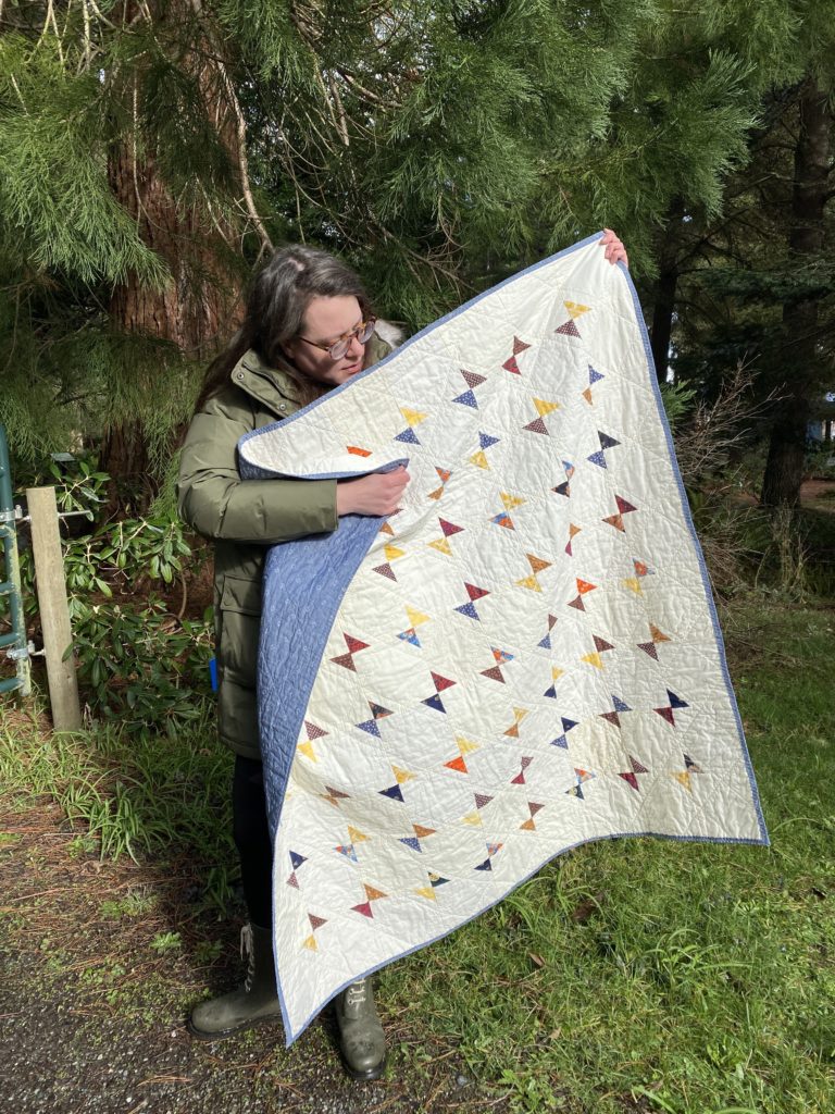 hourglass: a quilt with multiple shades and patterns of ivory fabrics as the background. Snowballed corners turn into hourglasses made from yellow, orange, blue and brown scraps. the backing and binding are blue chambray with small white anchors printed in rows. the square quilt is held by a femme in glasses, an olive-green coat and matching green boots.