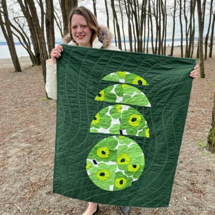 fem with long hair in white jacket holds a circular patterned green floral quilt with scalloped surface quilting